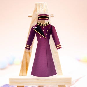 May include: A flat, acrylic-style decorative charm shaped like a long, deep purple coat with gold-colored buttons, a black lapel, and a matching pillbox hat. The piece is displayed on a small, light-colored wooden easel stand for vertical support.