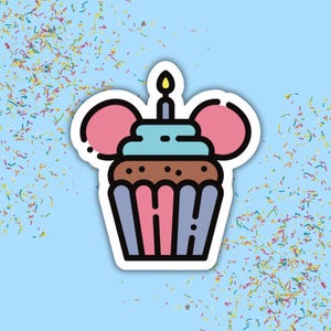 Op de afbeelding: Een sticker met een bruine cupcake met lichtblauwe glazuur, een enkele brandende kaars bovenop en twee grote roze ronde oren aan de zijkanten. De cupcake heeft een roze en lichtblauw gestreept papiertje op een lichtblauwe achtergrond.