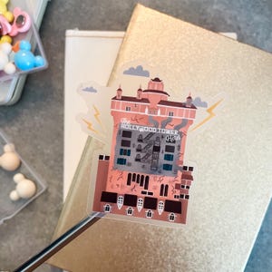 May include: A die-cut sticker featuring a stylized illustration of The Hollywood Tower Hotel. The building is rendered in shades of pink and brown with dark windows, a central fire escape, and yellow lightning bolts on the sides against a light sky.