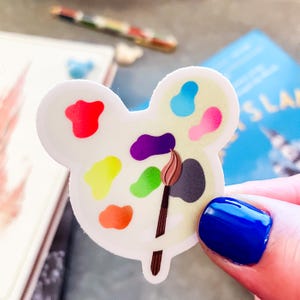 May include: A white sticker in the shape of a mouse head, featuring a colorful artist's palette with a paintbrush. The palette has red, yellow, orange, green, purple, blue, and pink paint splotches. The sticker is held by a hand with blue nail polish.