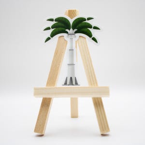 May include: A sticker featuring a stylized white tower with a green palm tree top, displayed on a small wooden tripod easel. The tower has a flared base and a segmented vertical structure, while the palm fronds are depicted in dark green.
