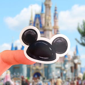 May include: A die-cut sticker featuring a glossy black Mickey Mouse ear hat with a white border. The design is a stylized illustration of the iconic headwear with rounded ears and a smooth, reflective surface, presented as a single adhesive decal.