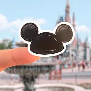Peut inclure: Un autocollant marron foncé en forme de tête de souris stylisée avec deux oreilles circulaires et une base arrondie. L'autocollant présente une finition brillante avec des reflets blancs et une fine bordure blanche, offrant un design graphique simple.