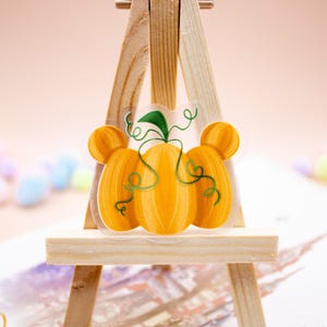 May include: A clear sticker featuring three orange pumpkins arranged in a cluster, with the two outer pumpkins shaped like mouse ears. The design includes a single green stem with curling vines extending across the orange pumpkin surfaces.