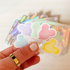 May include: A collection of die-cut stickers shaped like mouse-eared balloons in pastel shades of pink, yellow, blue, green, and purple. Each sticker features a white outline and a small balloon string, packaged inside a shimmering iridescent pouch.