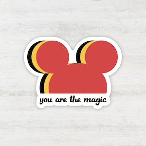 Puede incluir: Una pegatina troquelada que presenta una silueta estilizada de orejas de ratón rojas con sombras desplazadas en negro y amarillo. El texto "you are the magic" está impreso en una fuente cursiva negra en minúsculas debajo de la forma.