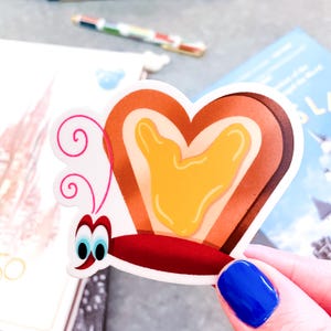 May include: A heart-shaped sticker featuring a cartoon butterfly with toast-like wings and a yellow honey center. The sticker has a red body, blue eyes, and pink antennae. The sticker is held in a hand with blue nail polish.