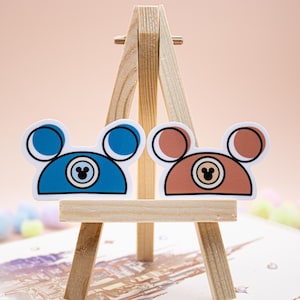 May include: Two die-cut stickers shaped like mouse ears, one in blue and one in brown. Each sticker features a central circular icon with a smaller mouse ear silhouette inside, outlined in black, displayed on a small wooden easel stand.