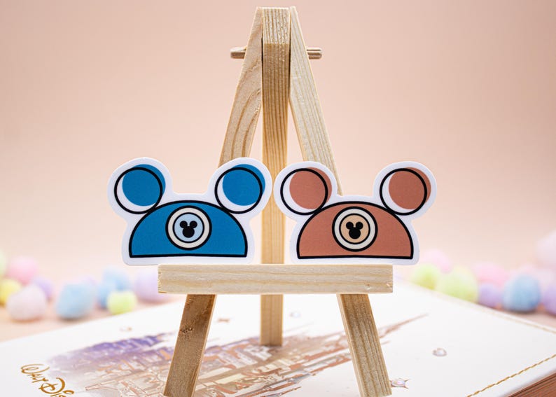May include: Two die-cut stickers shaped like mouse ears, one in blue and one in brown. Each sticker features a central circular icon with a smaller mouse ear silhouette inside, outlined in black, displayed on a small wooden easel stand.