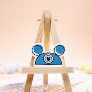 May include: A blue sticker shaped like a mouse ear hat with a white border. The design features two circular ears, a central circular emblem with a small mouse head silhouette inside, and a flat bottom edge, all displayed on a wooden easel.