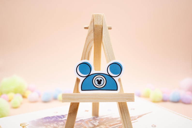May include: A blue sticker shaped like a mouse ear hat with a white border. The design features two circular ears, a central circular emblem with a small mouse head silhouette inside, and a flat bottom edge, all displayed on a wooden easel.