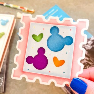 May include: A pink sticker with a postage stamp design. The sticker features a blue Mickey Mouse head, a purple Mickey Mouse head, and green and orange hearts. Small red and blue dots are scattered around the design.