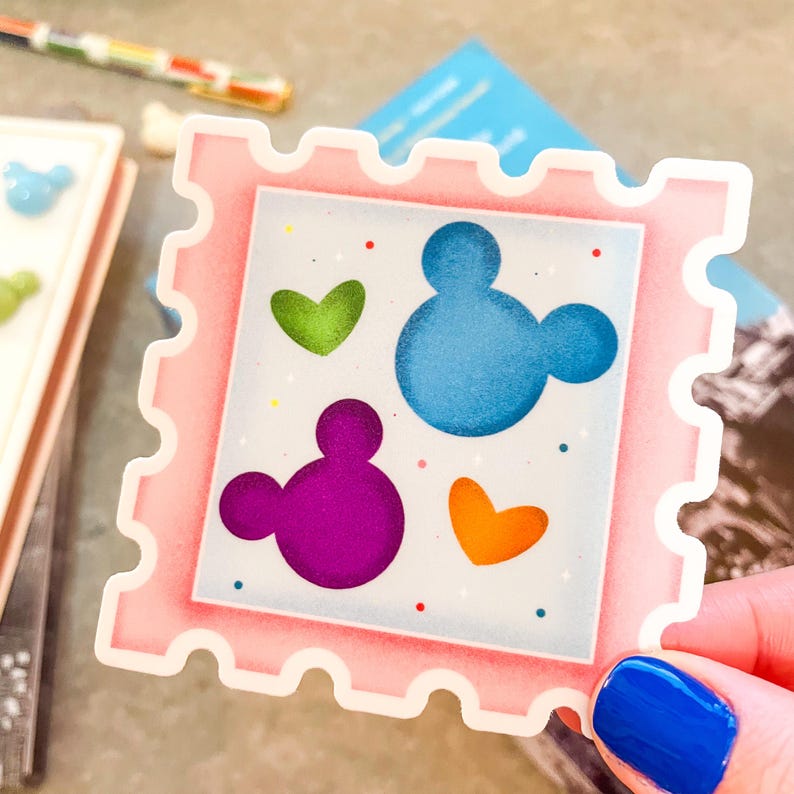 May include: A pink sticker with a postage stamp design. The sticker features a blue Mickey Mouse head, a purple Mickey Mouse head, and green and orange hearts. Small red and blue dots are scattered around the design.