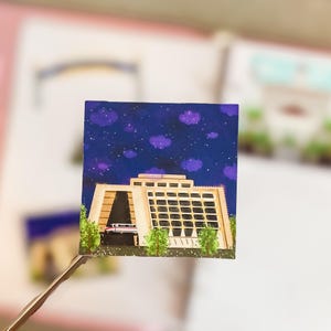 May include: A square sticker featuring a painting of a tan-colored building with a grid of windows and a monorail train emerging from a dark tunnel. The scene is set against a dark blue night sky filled with purple clouds and white stars.