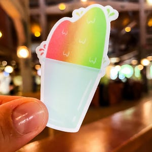 Può includere: Un adesivo fustellato raffigurante una granita colorata in un bicchiere trasparente. La granita presenta una sfumatura di rosso, arancione e verde brillante, decorata con piccole forme a 'w' bianche e accenti verdi. Il bicchiere è azzurro.