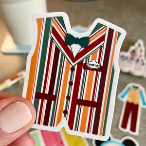 May include: A sticker featuring a colorful striped vest design with a green bow tie and the text "Dapper Dan". The vest has vertical stripes in orange, green, and burgundy.