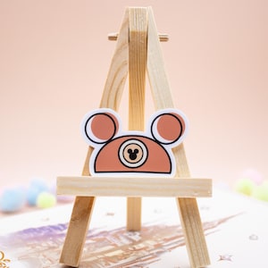 May include: A sticker featuring a stylized brown bear head with rounded ears and a central circular emblem containing a small black mouse silhouette. The sticker is displayed on a small wooden tripod easel, showcasing a clean design with bold outlines.