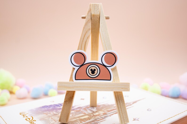 May include: A sticker featuring a stylized brown bear head with rounded ears and a central circular emblem containing a small black mouse silhouette. The sticker is displayed on a small wooden tripod easel, showcasing a clean design with bold outlines.