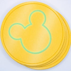 May include: A stack of yellow coasters featuring a light green outline of a Mickey Mouse head. The coasters are circular and have a slightly textured surface. The design is simple and recognisable.