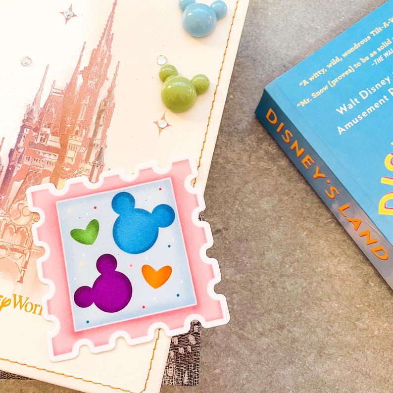 May include: A light-colored journal with a pink postage stamp sticker featuring Mickey Mouse ears and hearts in blue, green, purple, and orange. Also, a blue book titled "Disney's Land" and Mickey Mouse-shaped embellishments.