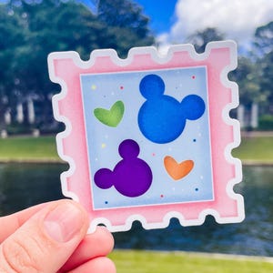 May include: A pink sticker with a postage stamp design. The sticker features a light blue background with a blue Mickey Mouse head, a purple Mickey Mouse head, and green and orange hearts. The sticker is held up by a hand.