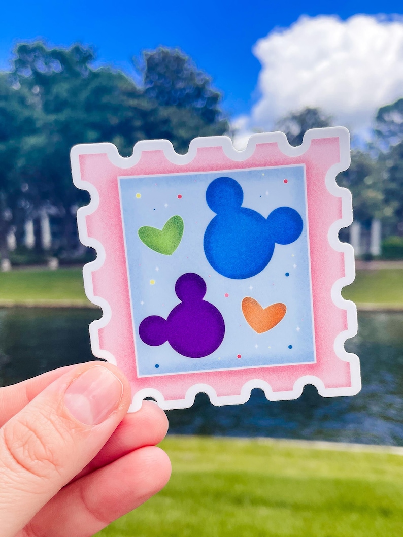 May include: A pink sticker with a postage stamp design. The sticker features a light blue background with a blue Mickey Mouse head, a purple Mickey Mouse head, and green and orange hearts. The sticker is held up by a hand.