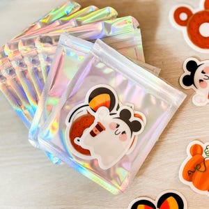 Puede incluir: Una pila de bolsas holográficas e iridiscentes resellables que contienen pegatinas troqueladas. La pegatina visible muestra un fantasma blanco con orejas de ratón negras sosteniendo una taza de café marrón, junto a una pegatina naranja de donut.