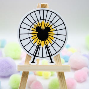 Könnte beinhalten: Ein runder Aufkleber mit einer schwarzen Mickey Mouse Silhouette in der Mitte, umgeben von einem gelb-weißen Design, das einer Sonnenblume ähnelt. Der Aufkleber wird auf einer kleinen Holzstaffelei mit bunten Pompons im Hintergrund präsentiert.
