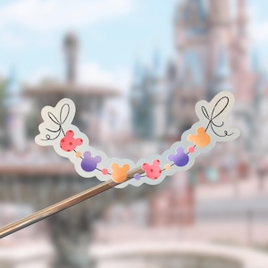 May include: A decorative sticker featuring a curved string of colorful Mickey Mouse-shaped icons in shades of red, purple, and orange, accented with small dots and tied at both ends with black line-art bows, presented on a clear backing.