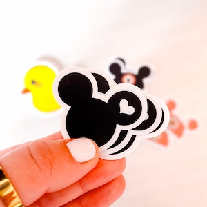 May include: A stack of black die-cut stickers shaped like a mouse head silhouette with a small white heart on the right ear. In the background, there are additional stickers featuring a yellow duck and a black bear character design.