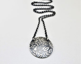 May include: A silver necklace with a round pendant featuring a detailed floral design. The pendant is textured and has a black oxidized finish.