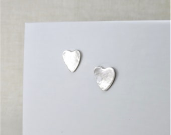 May include: Two silver heart-shaped stud earrings. The earrings have a hammered texture and are displayed on a white surface. The hearts are slightly irregular in shape, adding to their charm. The background is a soft gray.