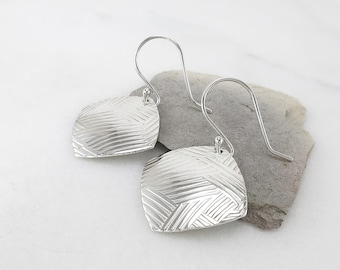 May include: A pair of silver earrings with a geometric design. The earrings are made of textured metal and have a square shape.