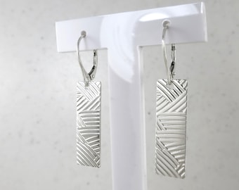 May include: Silver rectangular earrings with a textured, woven-like pattern. The earrings hang from a lever back closure. The earrings are displayed on a white stand against a neutral background.
