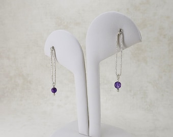 May include: Silver-tone earrings with a textured, elongated design. Each earring features a small, round, purple gemstone at the bottom. The earrings are displayed on a white jewelry stand against a neutral background.