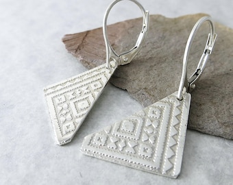 May include: A pair of silver-toned earrings. Each earring features a geometric design with intricate patterns. The earrings have a triangular shape with a lever-back closure. The earrings are displayed on a neutral-colored surface.