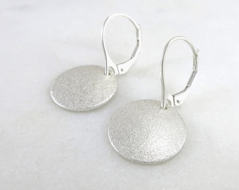 May include: A pair of silver-colored disc earrings with lever-back closures. The earrings have a textured, matte finish and a circular shape. The earrings are displayed on a white surface.
