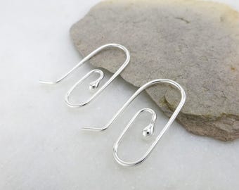 May include: A pair of silver-colored, wire-frame earrings. Each earring features a curved hook that transitions into a rectangular shape with a small, round bead at the bottom. The earrings are displayed on a light gray stone surface.