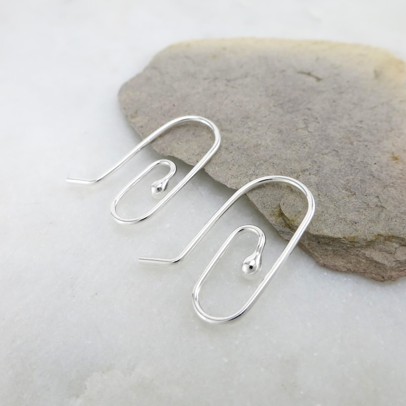 May include: A pair of silver-colored, wire-frame earrings. Each earring features a curved hook that transitions into a rectangular shape with a small, round bead at the bottom. The earrings are displayed on a light gray stone surface.