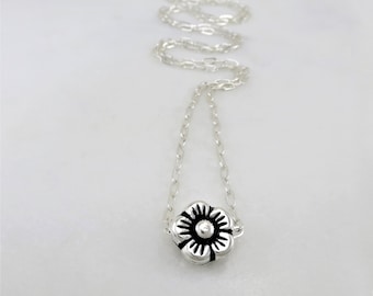 May include: A silver chain necklace with a small silver flower pendant. The flower has a black center and five petals.