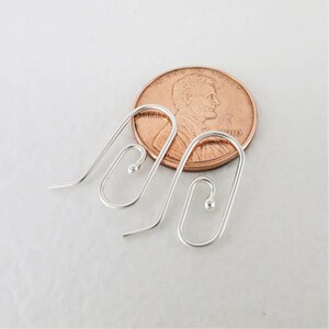 May include: Two silver-colored earring hooks with a curved design and a small ball at the end. A copper penny is next to the earrings for size comparison. The earrings are on a white surface.