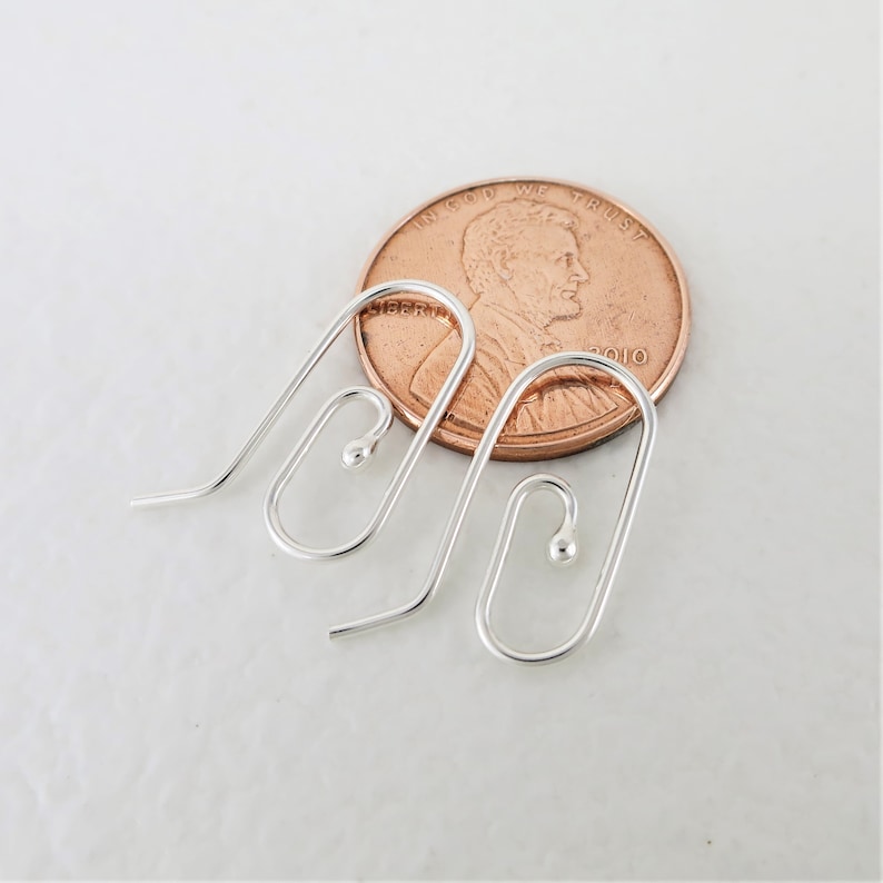 May include: Two silver-colored earring hooks with a curved design and a small ball at the end. A copper penny is next to the earrings for size comparison. The earrings are on a white surface.