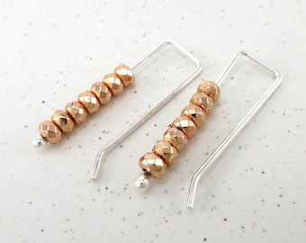 May include: A pair of U-shaped silver-toned earrings, each featuring a row of six faceted rose gold-colored beads. The earrings have a simple wire design with a small silver-toned bead at the end of the wire to secure the faceted beads in place.