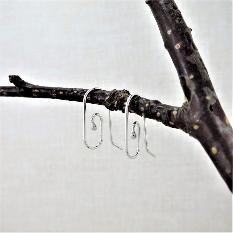 May include: A pair of silver-colored, minimalist earrings with a unique, curved design. The earrings are displayed on a dark brown tree branch against a white background. The earrings have a small, round detail at the top of the curved design.