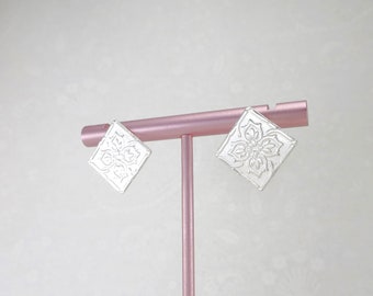 May include: A pair of silver earrings with a square design featuring a white floral pattern.
