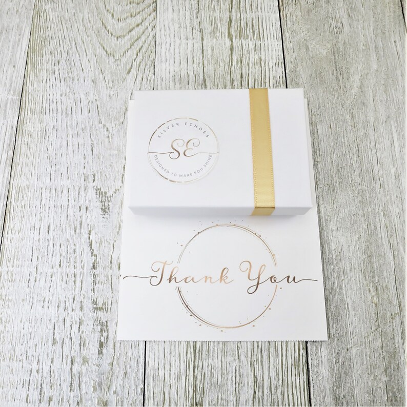 May include: A white box with a gold ribbon and a logo that says "Silver Echoes Designed to Make You Shine" and a white card with a gold circle and the text "Thank You".
