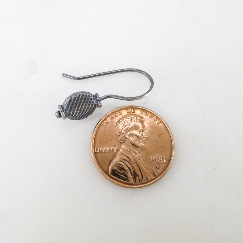 May include: A silver earring with a textured, round design. The earring is shown next to a US penny for size comparison.