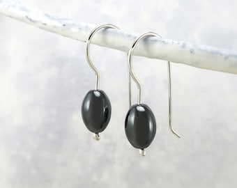May include: A pair of dark gray oval earrings with silver-colored hooks. The earrings are hanging from a white branch against a soft, blurred background. The earrings have a glossy finish and a small silver bead at the bottom.