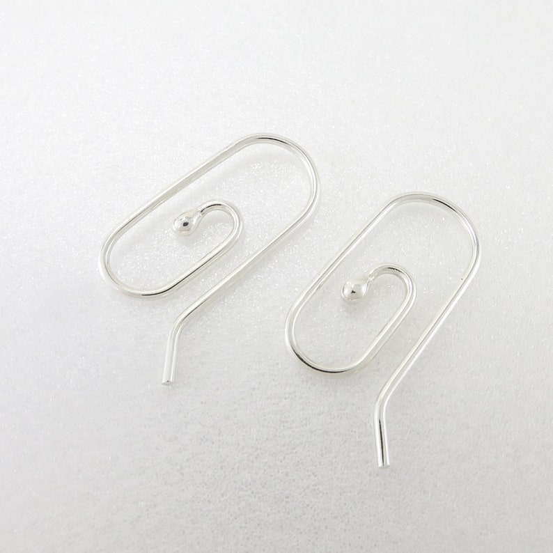 May include: A pair of silver-colored earrings. Each earring features a unique design with a small ball at the top of a spiral shape, connected to a curved hook. The earrings are displayed on a white surface.