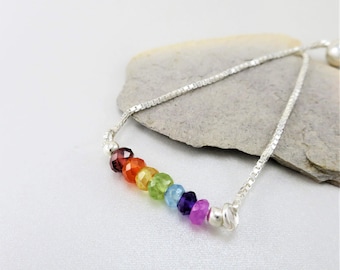 May include: A silver chain bracelet with a rainbow of gemstones. The bracelet has a sliding clasp and is made with red, orange, yellow, green, blue, purple, and pink gemstones.